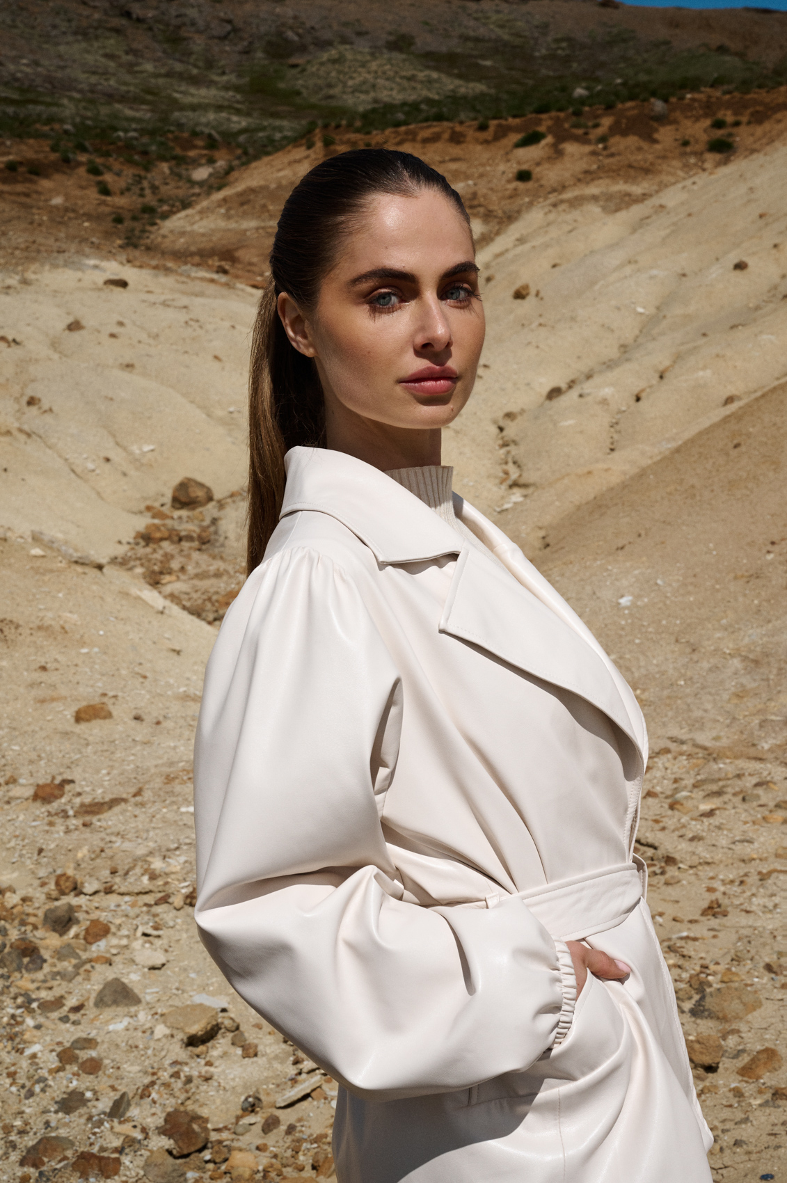 Fashion model dat een witte, leren jas draagt en voor een mooi landschap in IJsland staat.