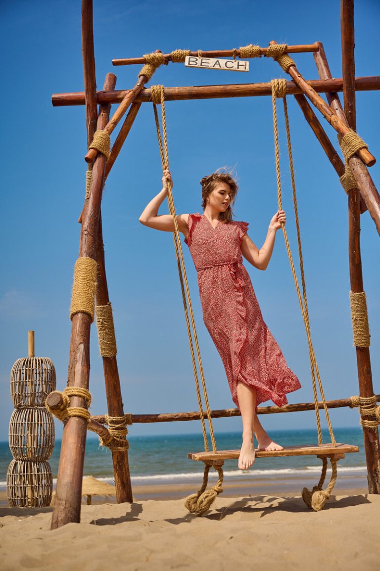 Een model dat op een schommel staat in een rode jurk. In de achtergrond zie je de zee.