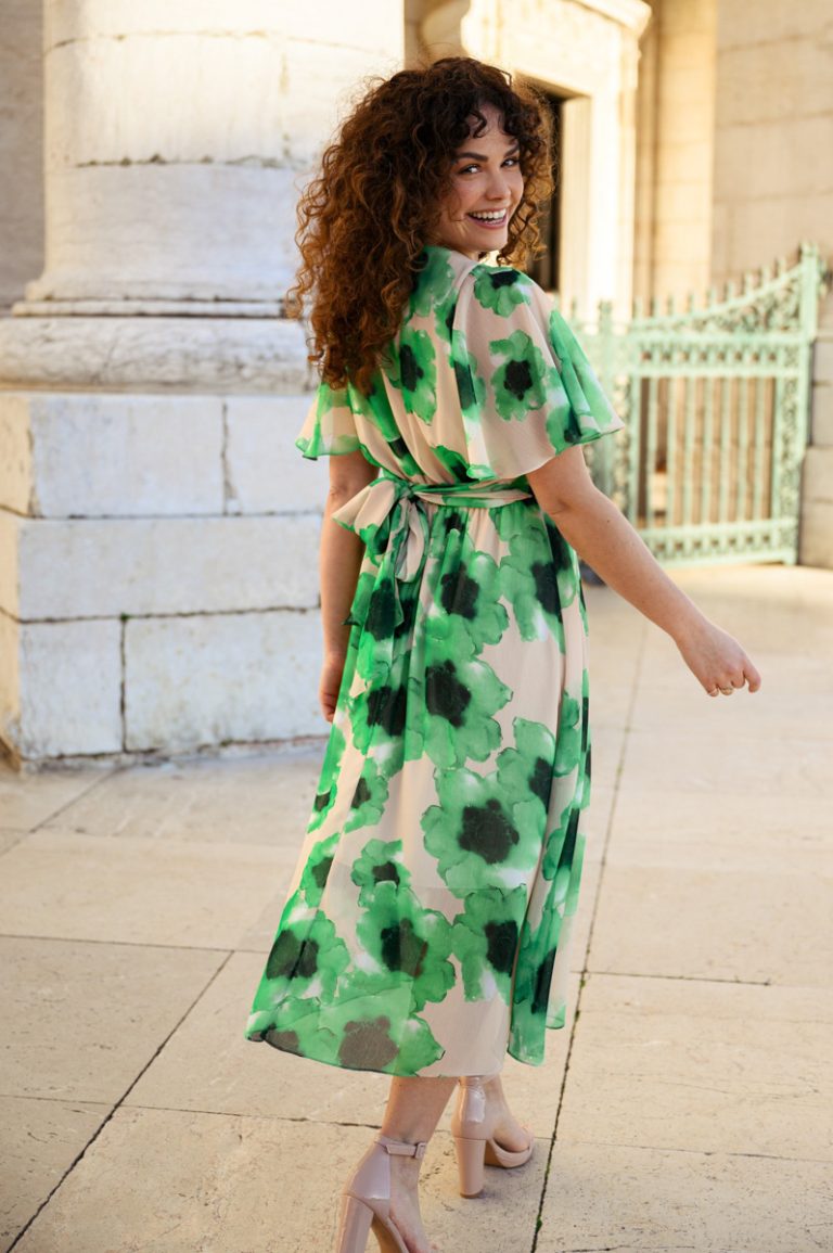 Model loopt naar voren en kijkt lachend over haar schouder in groene jurk voor een wit gebouw.