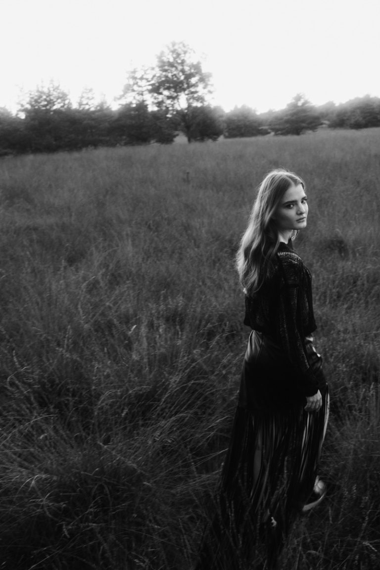 Model loopt door heide in Nederland in zwarte jurk en kijkt over haar schouder in de camera