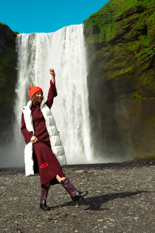 Lachend model staat met hand omhoog voor een grote waterval in IJsland met rode muts.